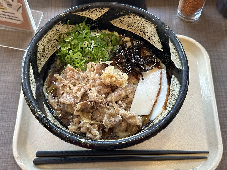 黒い器の肉ぶっかけうどん。ベージュのトレーに箸が添えられている。