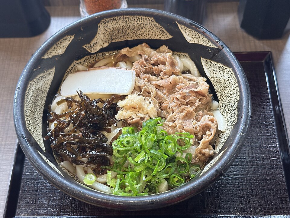 黒い器に盛られたうどんに、肉、ねぎ、昆布、天かす、かまぼこがのった肉ぶっかけ