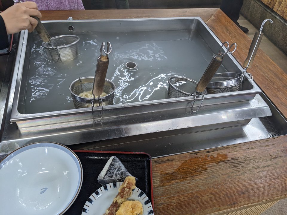 名玄の店内で、網かごに入れたうどんを温める湯槽。手前に天ぷらとおにぎりが写る。2023年2月撮影。